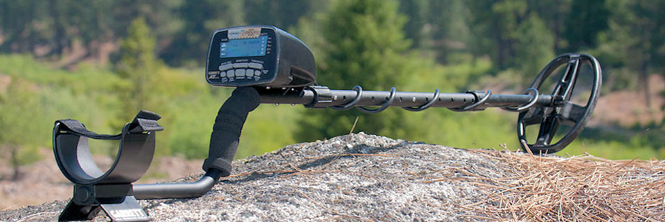 A metal detector with digital display resting on a rock outdoors, with green trees in the background, in soft daylight